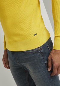 Żółty sweter z długim rękawem o prążkowanej strukturze, dopasowany fason, w połączeniu z ciemnoniebieskimi dżinsami. Zawiera małą metkę marki z boku.
