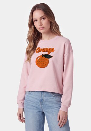 Giovane donna con lunghi capelli mossi che indossa una felpa rosa con un frutto arancione e la parola "Orange" in lettere arancioni.