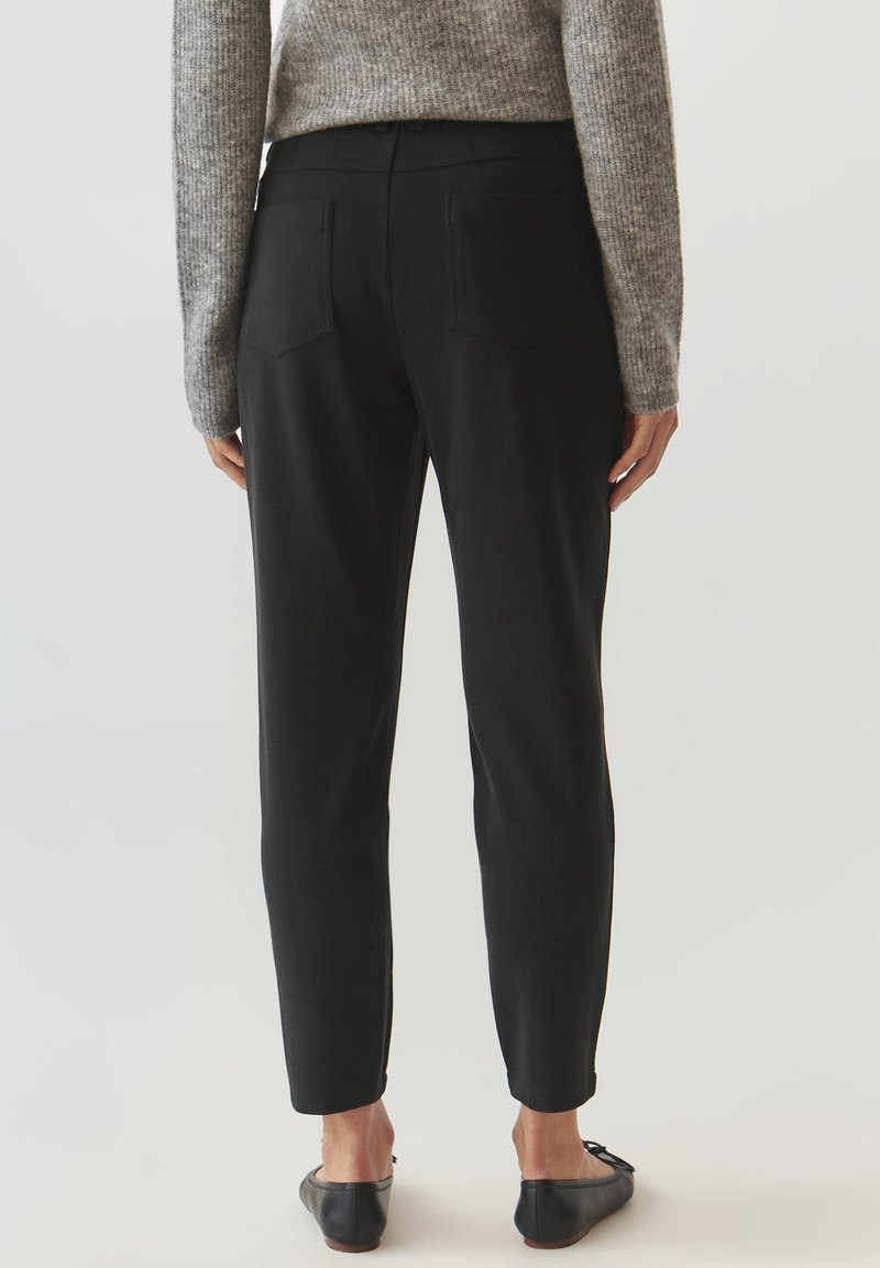 Czarne dopasowane spodnie o gładkiej fakturze, z dwoma tylnymi kieszeniami i zwężanym krojem. W połączeniu z szarym wełnianym swetrem.
