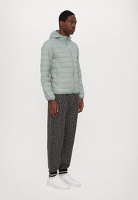Lys grøn puffjakke med hætte og quiltet tekstur, parret med sorte mønstrede joggingbukser og hvide sneakers.