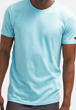 Homme portant un t-shirt bleu clair à manches courtes et un pantalon foncé, montré des épaules aux hanches sur fond blanc.