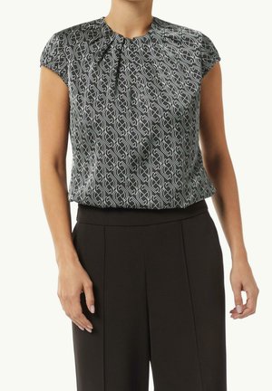 Femme portant un pantalon noir taille haute et un chemisier noir à manches courtes avec un motif géométrique blanc, debout les bras détendus.