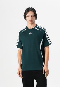 Mørk grønn treningst-skjorte med korte ermer, hvite sidelapper og tre hvite striper på hver skulder. Har et brodert logo.