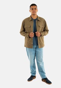 Camicia marrone con bottoni e due tasche sul petto, abbinata a una maglietta blu e jeans azzurri chiari. Sneakers nere con dettagli arancioni.