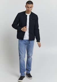 Jachetă bomber matlasată de culoare bleumarin, cu un buzunar cu fermoar, guler și manșete ribbate, asortată cu blugi deschiși la culoare, șifonați și adidași negri.