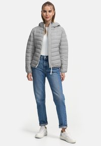 Grijze pufferjas met een capuchon, voorzien van een gewatteerd ontwerp en witte accenten. Gecombineerd met hoge blauwe jeans en witte sneakers.