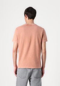 Tricou cu mânecă scurtă, de culoare coral estompată, cu guler rotund și croială lejeră, asortat cu blugi gri deschis, văzut din spate.