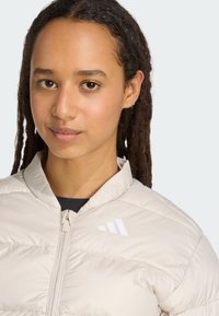 Young person with dreadlocks wears a light beige puffer jacket, looking directly at the viewer with a neutral expression.