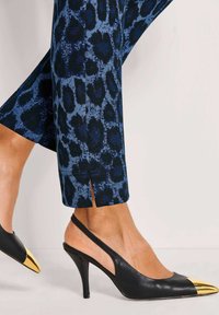 Benen in blauwe en zwarte panterprint broeken, gecombineerd met zwarte slingback hakken met metallic gouden puntneus.