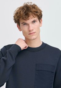 Donker marineblauwe crewneck sweatshirt met lange mouwen, voorzien van een borstzak en een textuur. Glad afwerkingspunt, eenvoudig ontwerp en geen graphics.