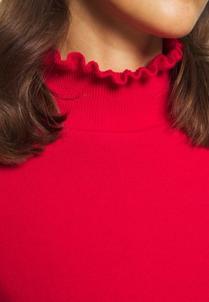 Nærbillede af en kvinde iført en lys rød ribstrikket rullekravesweater med en flæsekanter og brunt hår, der er delvist synligt.