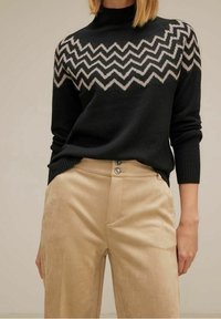 Kvinna bär en svart tröja med beige zigzag-mönster på överkroppen, i kombination med högmidjade beige byxor med knappstängning.