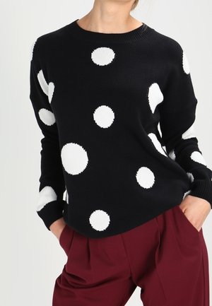 Pull noir à gros pois blancs associé à un pantalon plissé rouge foncé, mains dans les poches, debout devant un fond uni.