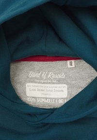 Teal puuvillaneuleen sisäosa harmaalla vuorauksella. Ominaisuuksiin kuuluu etiketti, jossa lukee "100 % puuvillaa (BIO)" ja "Global Organic Textile Standard."