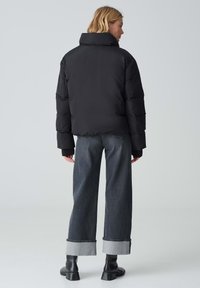 Giacca a vento nera con collo alto, caratterizzata da una vestibilità aderente e polsini a coste, indossata con jeans grigi larghi e stivaletti alla caviglia.