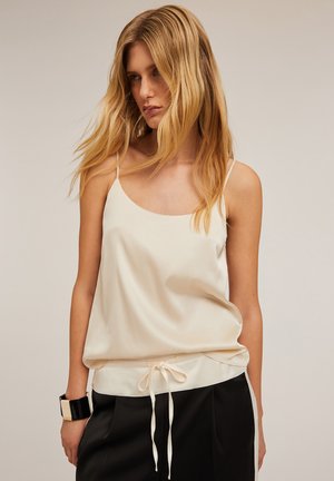 Satin cami i lys beige med tynde stropper, rundet udskæring, snoreliv og glat tekstur. Sættes sammen med sorte skræddersyede bukser.