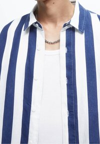 Niebiesko-biała w paski koszula z guzikami i kołnierzykiem, wykonana z lekkiego materiału i z widocznymi guzikami. Nałożona na białą koszulkę bez rękawów.