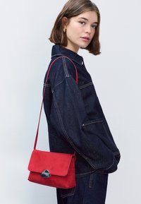 Červená semišová kabelka na rameno so zapínaním na chlopňu a striebornými kovaniami, nesená cez denimovú bundu. Kabelka je kompaktná a má obdĺžnikový tvar.