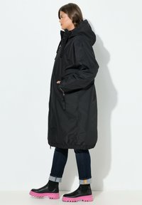 Abrigo negro impermeable con capucha, que cuenta con cordón ajustable, bolsillos laterales y dobladillo elástico. Combinado con botas negras hasta el tobillo y jeans oscuros.