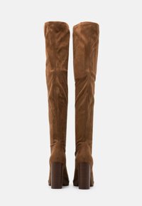 Brązowe kozaki za kolano z miękkiego zamszu, z wysokim, szerokim obcasem i zaokrąglonym noskiem. Szwy zdobiące tył. Teksturowany akcent na obcasie.