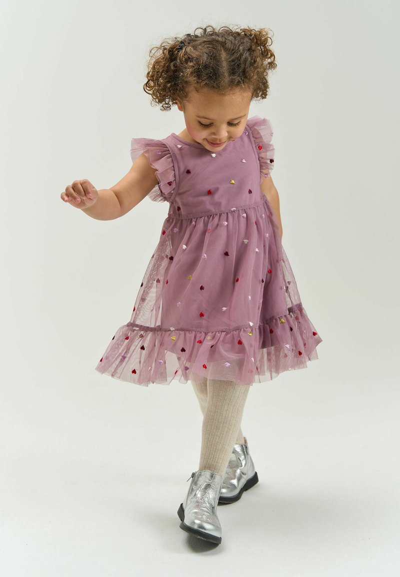 Jeune fille aux cheveux bouclés portant une robe rose à motifs de cœurs, des collants beiges et des bottes argentées, marchant avec un léger sourire sur un fond uni.