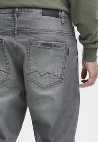 Jeans din denim gri au două buzunare spate cu un model distinctiv de cusătură în zigzag. Talia are un eticheta din piele neagră.