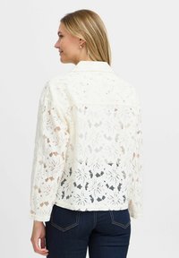 Laceblus i krämfärg med ett blommönster, som har krage och långa ärmar, delvis fodrad med en genomskinlig baksidesdesign.