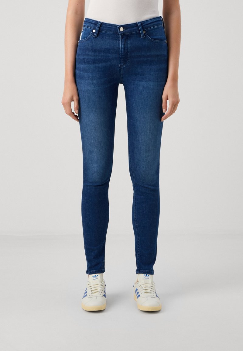 Jeans ajustados de denim oscuro con una textura suave, diseño de cinco bolsillos y trabillas para cinturón. Combinados con zapatillas blancas con rayas azules.