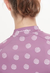 Lila Sportshirt mit hohem Kragen, das ein gepunktetes Muster in Weiß aufweist. Der Stoff wirkt glatt und hat eine taillierte Passform.