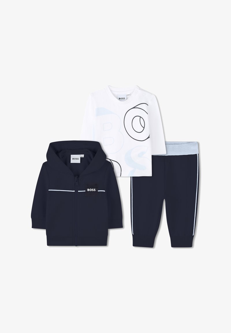 Felpa con zip blu navy con dettagli bianchi, camicia a maniche lunghe bianca con logo blu e pantaloni della tuta blu navy con vita azzurra chiara.