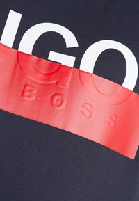Navy cotton t-paita, jossa on rohkea punainen vaakaraita, ja jossa on kohotettu valkoinen teksti "BOSS". Sujuva pinta, rento muotoilu.