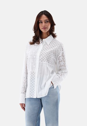 Camicia bianca con bottoni, caratterizzata da una fusione di motivi floreali ricamati e tagli a occhio. Maniche lunghe, colletto classico e vestibilità rilassata.