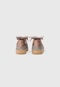 Zapatillas de deporte en rosa dorado brillante con parte superior brillante, acentos texturizados y un lazo. Presentan una suela de goma y un logotipo en el talón.