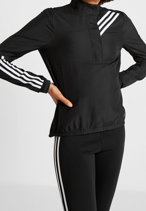 Person wearing a black sports jacket and leggings with three white stripes on sleeves, chest, and sides, standing with hands on hips.