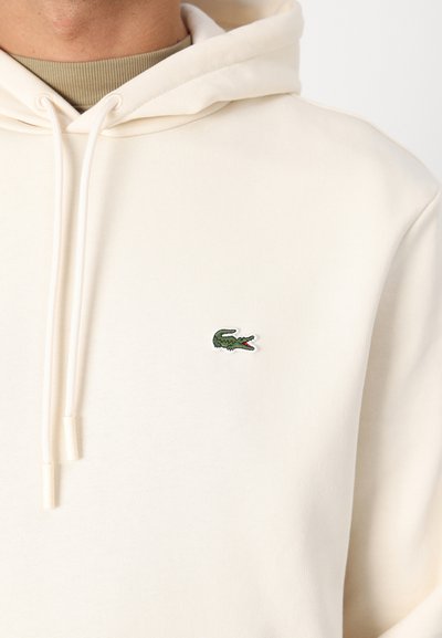 Sweatshirt crème en tissu doux, doté d'une poche kangourou à l'avant, de cordons de serrage et d'un petit logo crocodile vert sur la poitrine gauche.