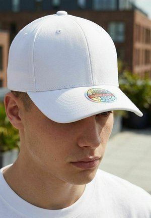 Young man wearing a plain white baseball cap with a round colorful label on the brim, outdoors in urban setting.