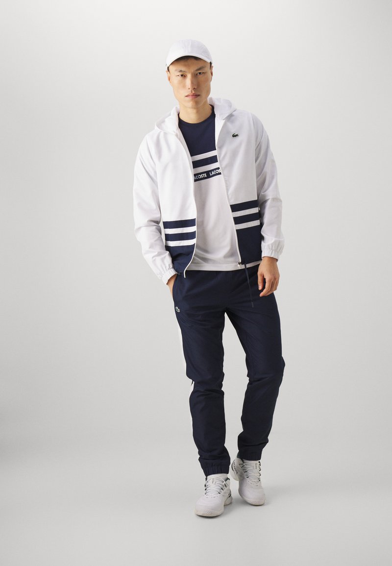 Conjunto atlético para hombres que presenta una chaqueta blanca con cremallera y rayas marinas, pantalones marinos y una gorra blanca, acentuados con un logo de Lacoste.