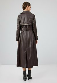 Donkerbruine leren trenchcoat met een ceintuur in de taille, grote kraag en een split aan de achterkant. Voorzien van een gouden kettingdetail op de achterkant.