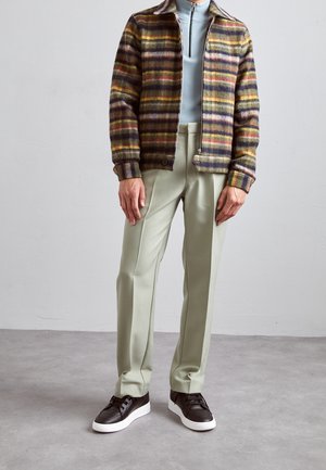 Uld ternet jakke med lynlås i brun, gul og grøn; lyseblå top med høj hals; lysegrønne bukser; sorte sneakers med hvide såler.