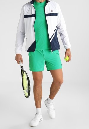 Männlicher Tennisspieler trägt eine weiße und marineblaue Jacke, grünes Hemd und Shorts, hält einen Tennisschläger in einer Hand und einen Tennisball in der anderen.