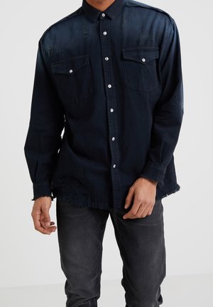 Uomo che indossa una camicia nera usurata con bottoni e tasche sul petto e jeans scuri su uno sfondo semplice.