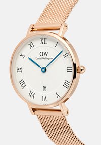 Reloj de oro rosa con un dial texturizado blanco, números romanos, manecillas azules y una pantalla de fecha. La correa de malla complementa el diseño.