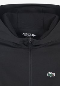 Μαύρη ζακέτα με φερμουάρ και διχτυωτή επένδυση, με μικρό πράσινο λογότυπο Lacoste. Λείο ύφασμα, αυτόνομη κουκούλα και εφαρμοστή σχεδίαση.