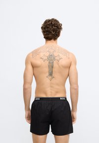 Čierne plavecké šortky s opaskom na zaväzovanie, ozdobené výrazným logom. Na chrbte sú viditeľné zložité tetovania na pokožke.