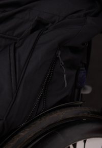Una tela negra con una textura suave presenta un bolsillo con cremallera en el lado de una silla de ruedas, adyacente a una rueda oscura y con estampado.