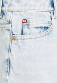 Ljust blå denimjeans med ett slitna mönster, synlig sömnad och ett röd tygmärke med "60" på framfickan.