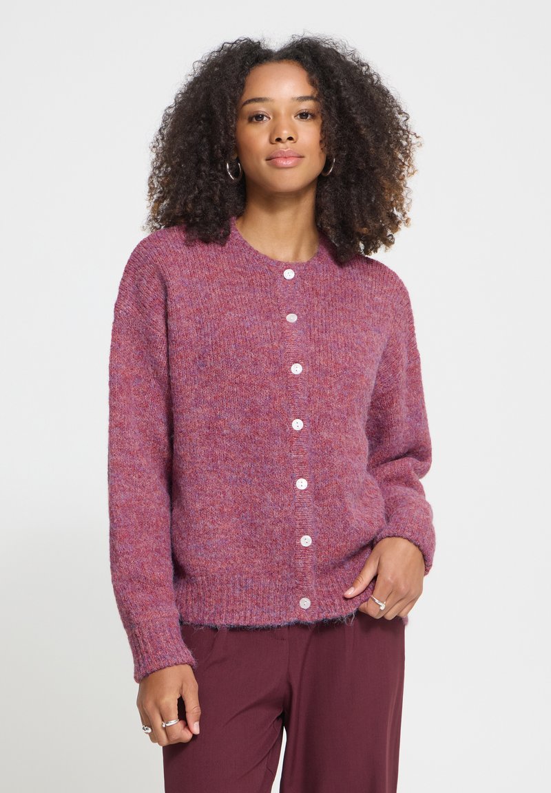Femme aux cheveux bouclés portant un cardigan violet boutonné et un pantalon bordeaux, debout avec une main dans la poche, devant un fond uni.