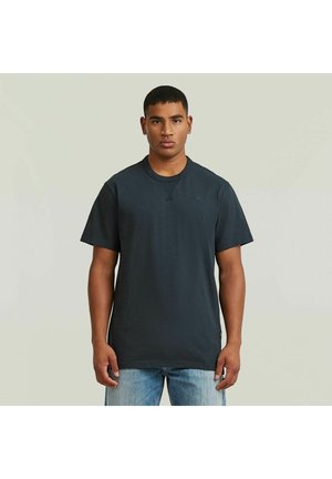 Ung mand, der står og vender fremad iført en ensfarvet mørkeblå t-shirt med rund hals og lyseblå jeans mod en neutral baggrund.