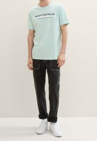 Mintová tričko s potiskem „DENIM TOM TAILOR“ v tmavých písmech, kombinované s černými džínami a bílými teniskami. Jednoduchý, ležérní design.