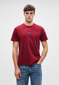 Burgundia puuvillane t-särk, lühivarrukatega ja ümar kaelus, mille rinnal on väike sinise ja valge logoga, koos siniste teksapükstega.